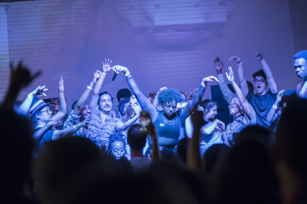 Joyful nighttime photo of a crowd of multiracial people surrounding a performer on stage.