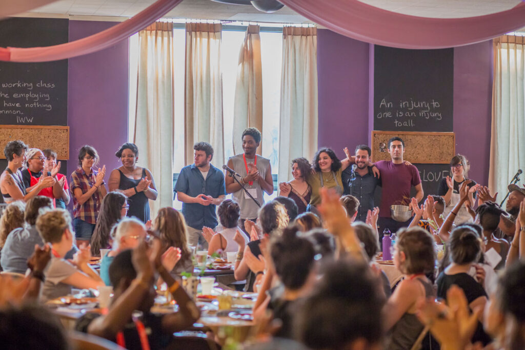 A celebratory meal at the AMC, with people at round tables clapping for a group standing shoulder to shoulder at the front of the room