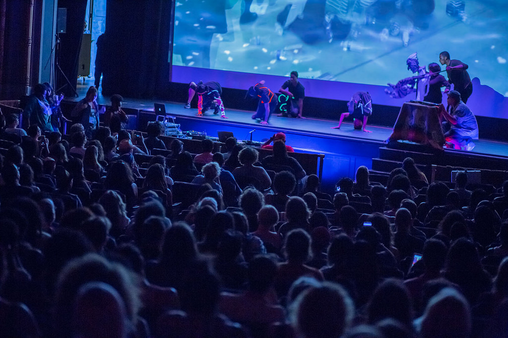Performance in an auditorium at the AMC with dark lighting and performers on stage wearig glowing stripes