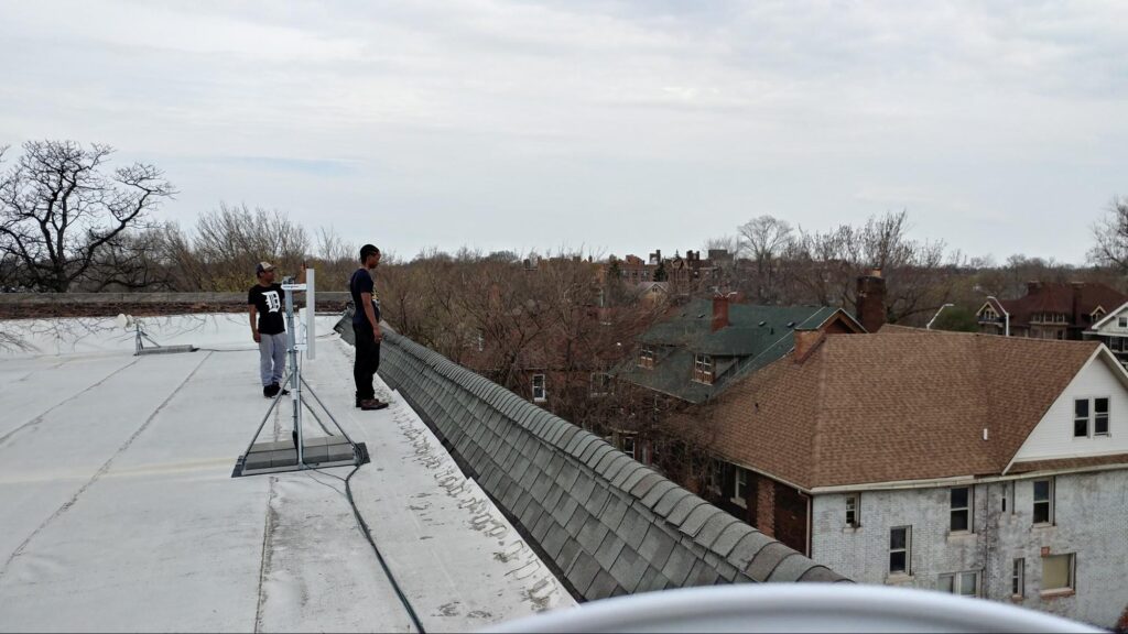 Two EII members on top of a commercial roof near a mesh network satellite, looking out over the residential skyline