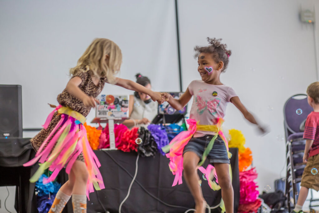 Two young children with facepaint and handmaid streamer skirts holding hands and dancing in front of a DJ table