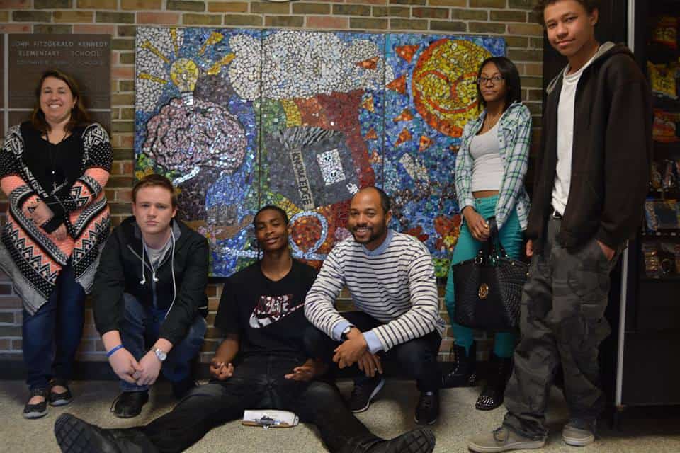 Four DFS participants, a female teacher, and DFS director standing and kneeling in front of their tile mural of a sun, rainbow, brain, lightbulb